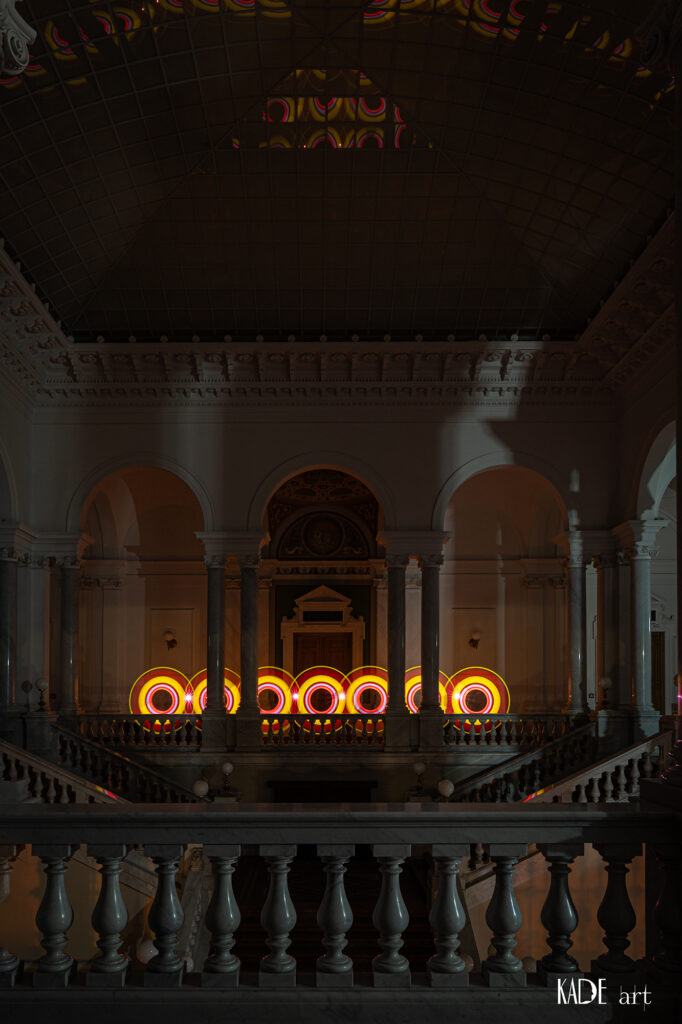 Lightpainting in der Bibliotheca Albertina. Zu sehen sind 7 Lichtkreise die perfekt neben einander gesetzt wurden. Teilweise werden die Kreise mit Strenchen unterbrochen. Die kreise haben von innen nach außen folgende Farben: Orange weiß rot gelb rot und wieder gelb.