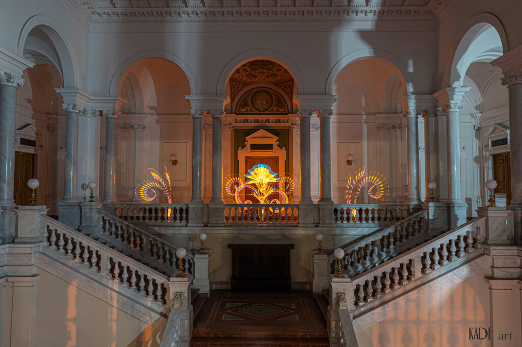 Lightpainting in der Bibliotheca Albertina Zu sehen sind 3 florale Ornamente im Foyer der Bibliothek. Die Farben sind gelb mit roten und blauen Elementen. Das Bild ist sehr symmetrisch und klar