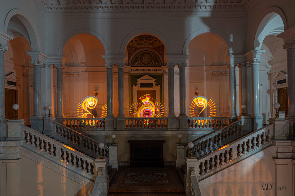 Lightpainting in der Bibliotheca Albertina zu sehen sind 3 florale Ornamente im Foyer der Bibliothek. Die Farben sind gelb mit roten und blauen Elementen. Das Bild ist sehr symmetrisch und klar
