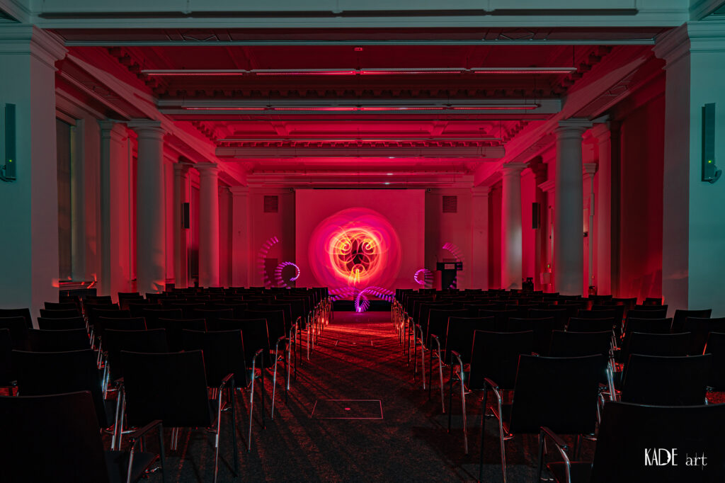 Zu sehen ist ein pinker Orb mit orangenem Zentrum. Er wurde auf der Bühne im Vortragsraum der BIbliotheca Albertina mit licht gemalt