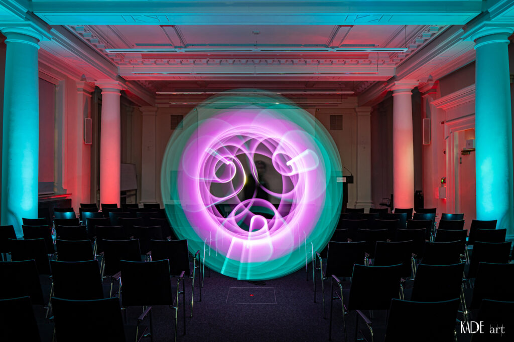 Zu sehen ist ein pink/türkies farbener Orb. Er wurde auf der Bühne im Vortragsraum der Bibliotheca Albertina mit licht gemalt